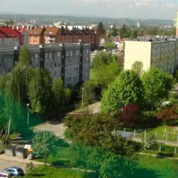 Panoramiczny widok z góry na osiedle mieszkaniowe z blokami o zróżnicowanej kolorystyce elewacji, zieleń drzew i trawników, parking z samochodami, oraz budynek szkoły w oddali.
