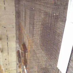 Siatka zbrojeniowa ułożona przy betonowej ścianie z widocznymi rurami i fragmentami rusztowania w tle, przygotowana do zalania betonem.