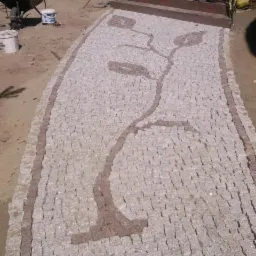 Ścieżka z jasnej kostki brukowej z motywem roślinnym w kolorze brązowym, prowadząca do schodów wejściowych do domu. W tle widoczny wózek budowlany i wiaderka z farbą.