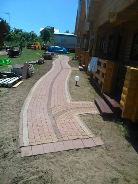 Kręta ścieżka z kostki brukowej w odcieniach beżu i brązu, prowadząca przez piaszczysty teren obok drewnianego domu, zakończona niewielkim placykiem i oświetlona małą, kulistą lampą.