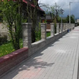 Długi, niski mur z kamiennych bloczków w odcieniach szarości i brązu, z kolumnami i podmurówką, oddzielający posesję od chodnika i drogi asfaltowej w słoneczny dzień.