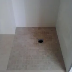 Ukończony brodzik wyłożony mozaiką w odcieniach beżu i brązu, z centralnie umieszczonym odpływem, otoczony gładkimi, beżowymi płytkami podłogowymi i białymi ścianami.