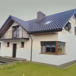 Dwupiętrowy dom z białymi ścianami, ciemną dachówką i balkonem z czarną balustradą, widok na elewację frontową i boczną, z zaznaczonymi elementami wykończenia.