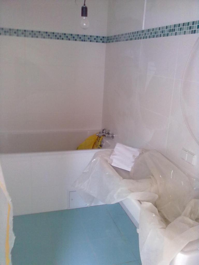 Niedokończona łazienka z niebieską podłogą, białą wanną i świeżo położonymi białymi płytkami na ścianach, zabezpieczonymi folią ochronną, widoczny fragment dekoracyjnego pasa płytek mozaikowych...