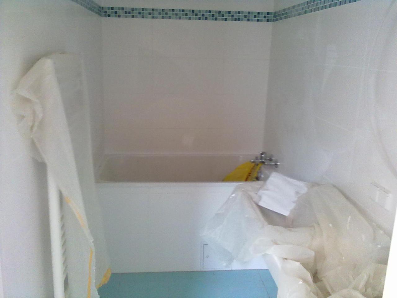 Łazienka w trakcie remontu: białe płytki ścienne z dekoracyjnym paskiem mozaiki, zabezpieczona folią wanna, niebieska podłoga.