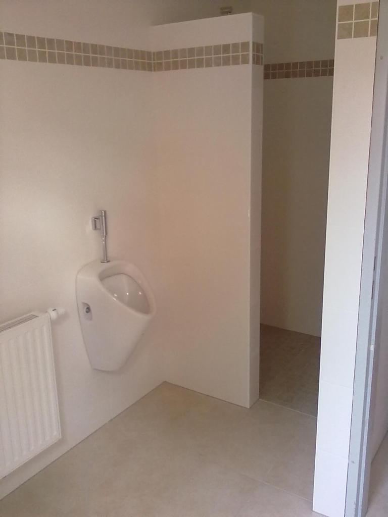 Biała pisuar wiszący na ścianie w toalecie wykończonej jasnymi płytkami, z dekoracyjnym pasem mozaiki u góry.