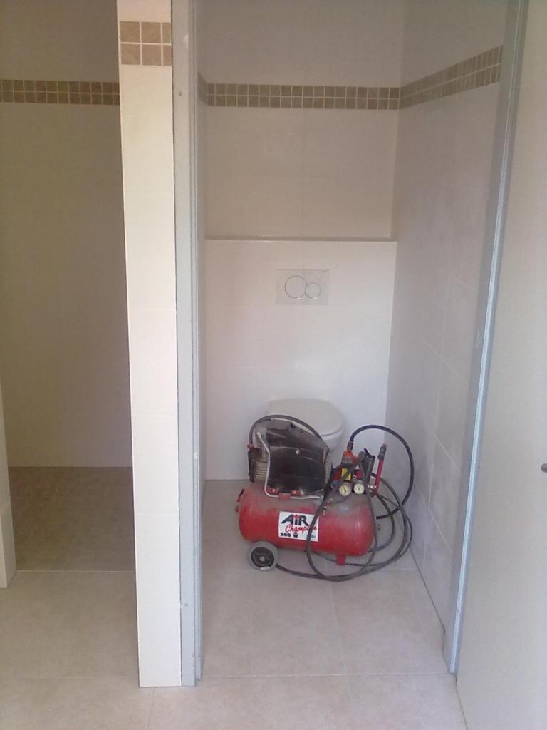 Czerwony kompresor Air Champ 3000 ustawiony w wykafelkowanej wnęce łazienki z zamontowaną toaletą, widoczne płytki i przyciski spłuczki.