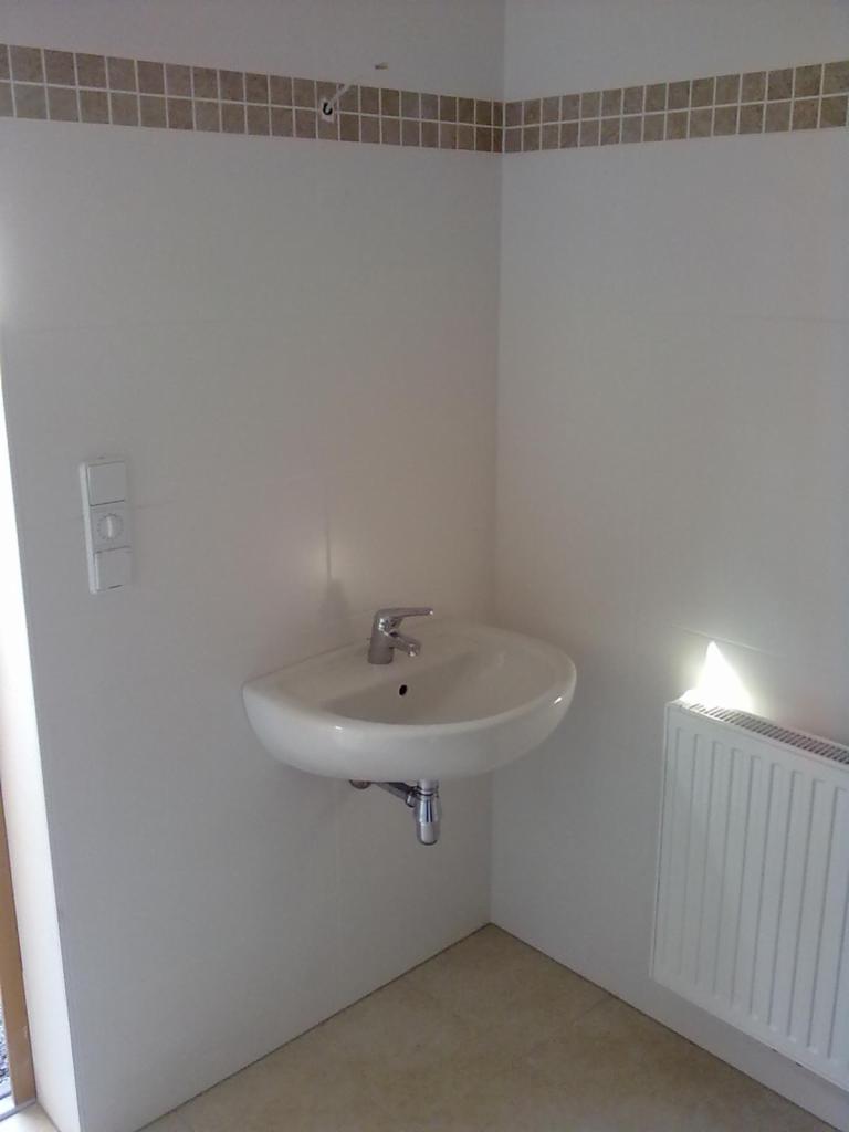 Mała umywalka zamontowana na białej ścianie w narożniku łazienki, nad nią pasek dekoracyjnych płytek, obok grzejnik.