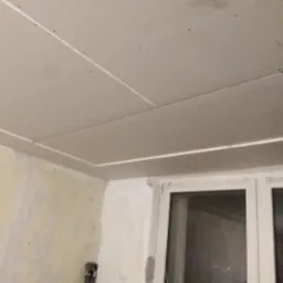 Częściowo zamontowana płyta kartonowo-gipsowa na suficie, widoczne łączenia, ściany w trakcie przygotowania, okno z białymi ramami.