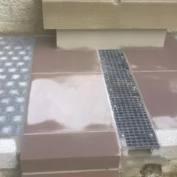 Ułożona kostka brukowa i płyty chodnikowe w odcieniach szarości i brązu, z metalową kratką odpływową pośrodku, przy beżowej ścianie z kamienia.