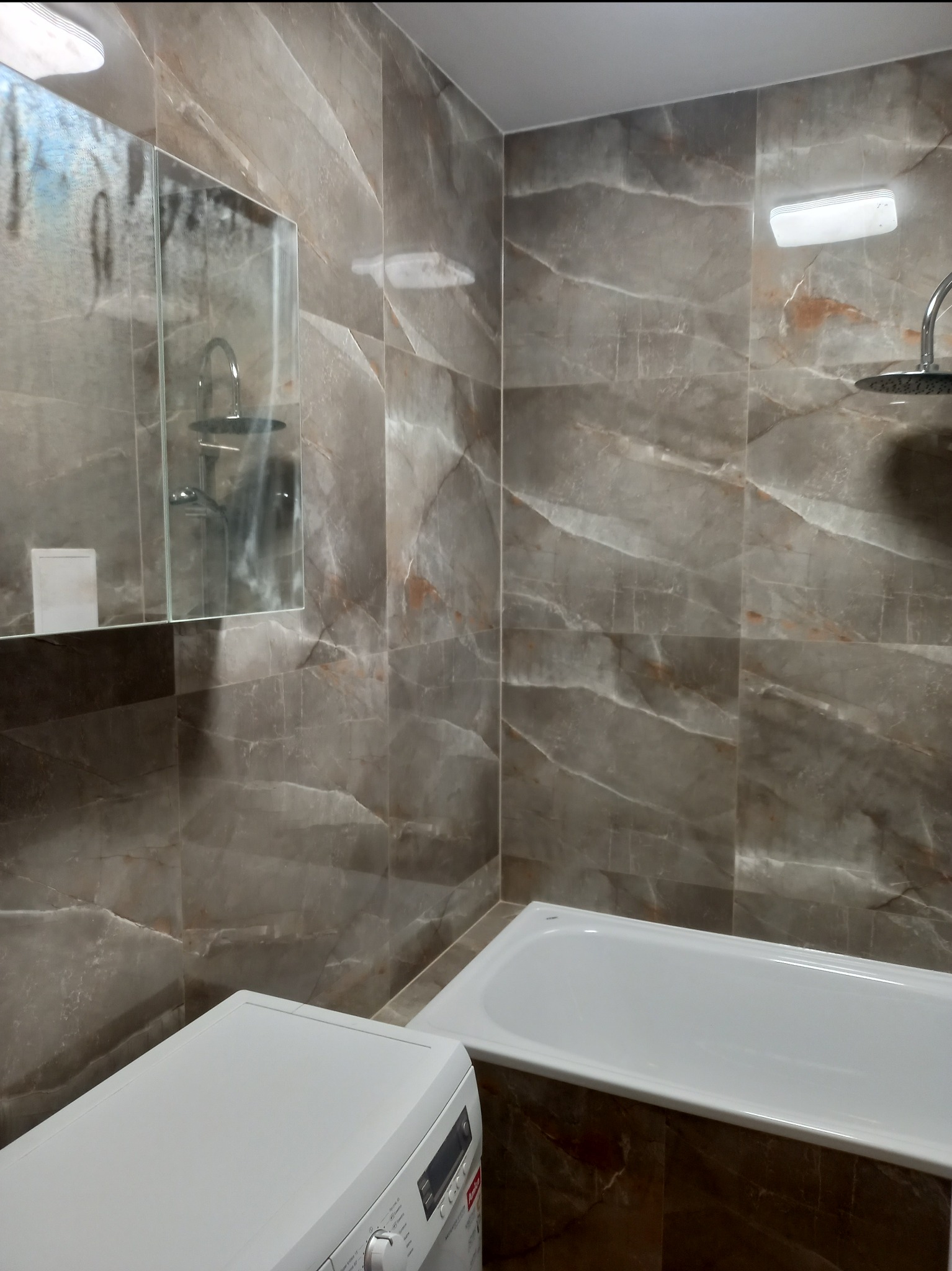 Wykończona łazienka z płytkami imitującymi marmur, białą wanną i pralką w dolnym rogu kadru. W lustrze odbija się armatura.