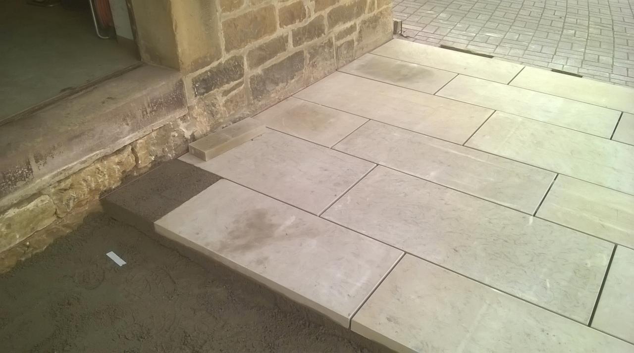 Układanie jasnych, prostokątnych płyt kamiennych na zaprawie piaskowo-cementowej przy wejściu do budynku z kamienia.