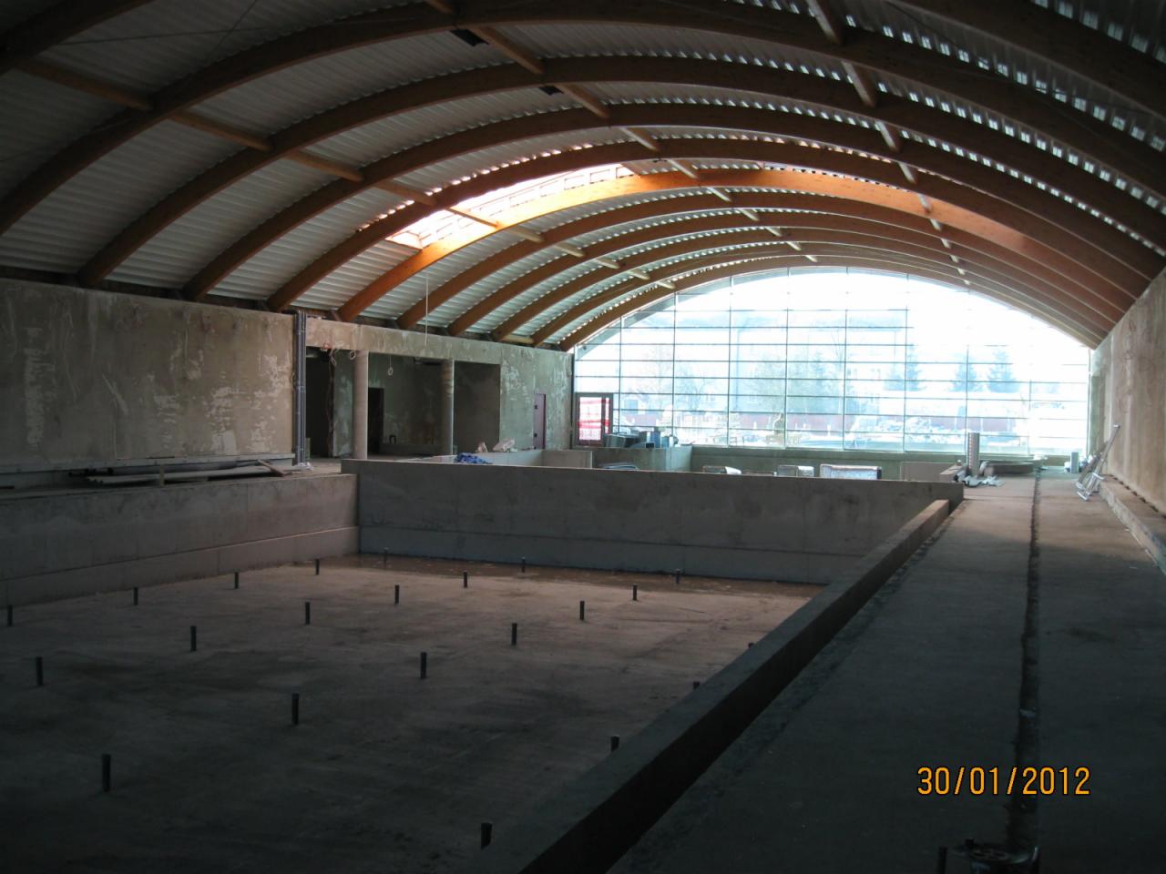 Wnętrze w trakcie budowy z widocznym szkieletem niecki basenowej, betonowymi ścianami i drewnianą konstrukcją dachu z oknami w tle.