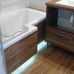 ABV Zbigniew Spławski - Nowoczesna łazienka z wanną obudowaną panelami imitującymi drewno, podświetleniem LED i szafką pod umywalkę z drewna w podobnym odcieniu, na podłodze płytki imitujące drewno.