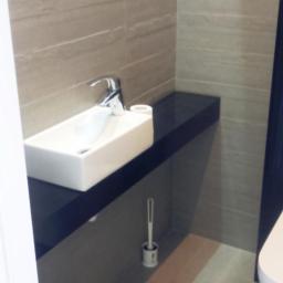 ABV Zbigniew Spławski - Nowoczesna umywalka nablatowa o prostokątnym kształcie, z chromowaną baterią, umieszczona na czarnym blacie w łazience wykończonej płytkami w odcieniach beżu i szarości, widoczny fragment toalety.