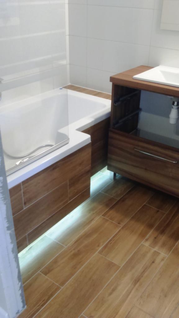 Nowoczesna łazienka z wanną obudowaną panelami imitującymi drewno, podświetleniem LED i szafką pod umywalkę z drewna w podobnym odcieniu, na podłodze płytki imitujące drewno.