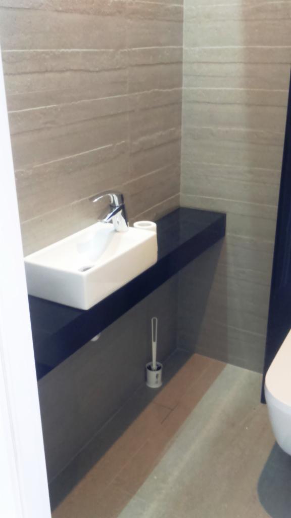 Nowoczesna umywalka nablatowa o prostokątnym kształcie, z chromowaną baterią, umieszczona na czarnym blacie w łazience wykończonej płytkami w odcieniach beżu i szarości, widoczny fragment toalety.