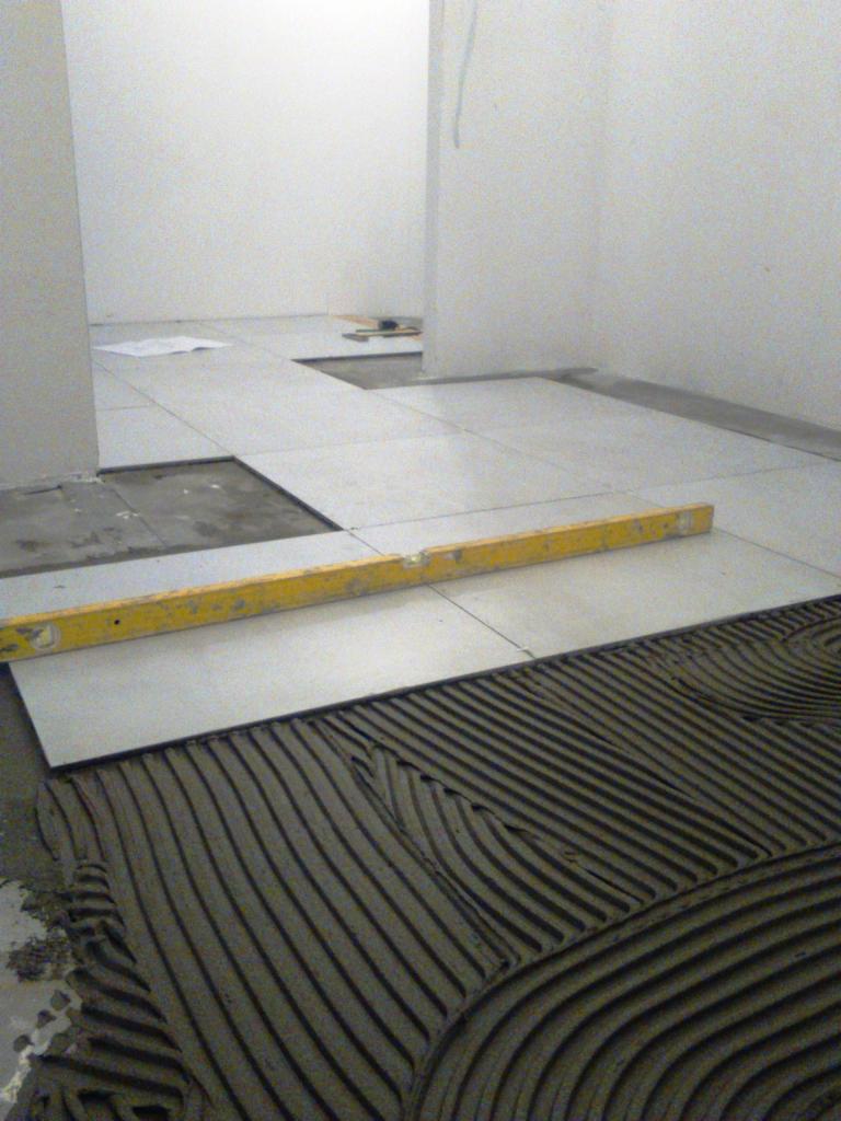 Układanie jasnych płytek podłogowych w łazience, widoczna warstwa kleju z charakterystycznymi rowkami, poziomica na płytce, surowe białe ściany w tle.