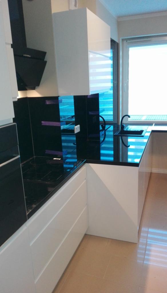 Nowoczesna kuchnia z białymi szafkami i czarnym blatem, odbijającym światło z okna, z widoczną płytą indukcyjną i minimalistycznym okapem.