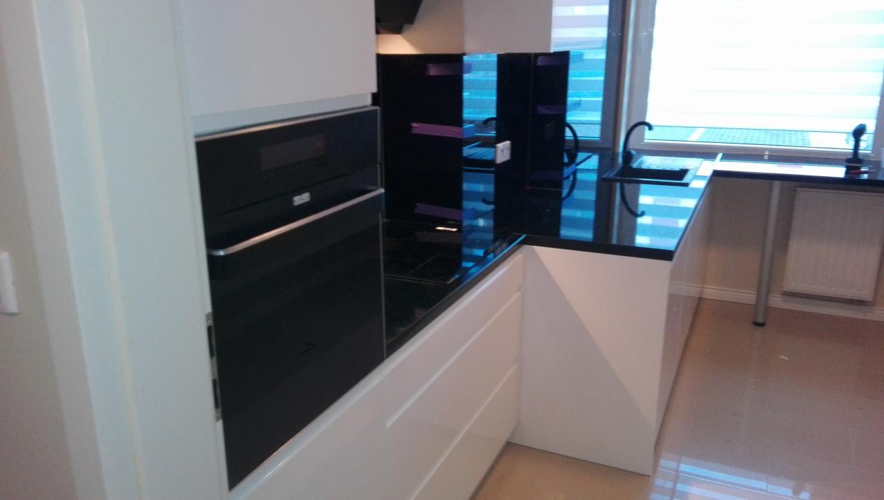 Nowoczesna kuchnia z białymi szafkami, czarnym blatem i szklanym panelem nad blatem z fioletowymi akcentami, widoczny piekarnik w zabudowie i zlewozmywak pod oknem z żaluzjami.