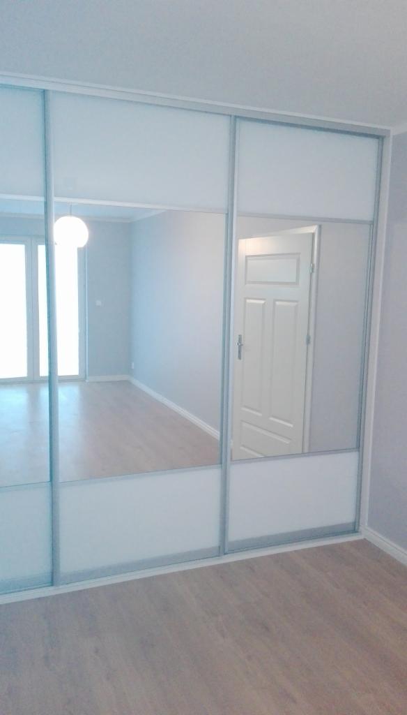 Biała szafa wnękowa z przesuwnymi drzwiami lustrzanymi, odbijająca wnętrze pokoju z drewnianą podłogą i białymi drzwiami.