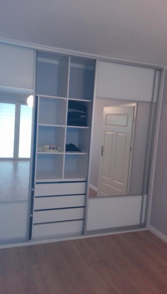 Biała szafa przesuwna z lustrem i otwartymi półkami, widoczne ubrania na półkach, drewniana podłoga.