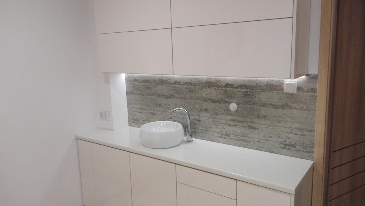 Biała, okrągła umywalka nablatowa z chromowaną baterią na blacie z szafkami w odcieniach bieli. Nad blatem panel dekoracyjny imitujący beton, podświetlony od góry. Widoczny fragment drewnianych drzwi.