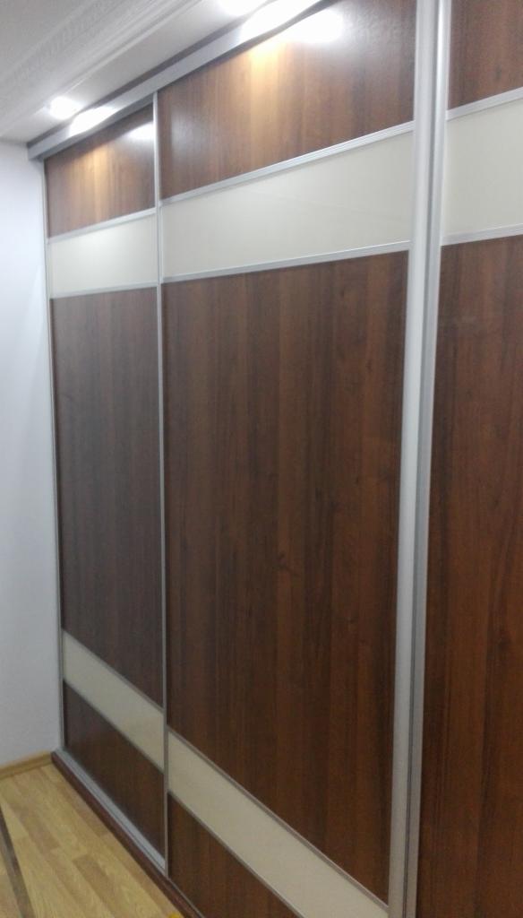 Trzydrzwiowa szafa przesuwna z ciemnego drewna z poziomymi wstawkami w kolorze kremowym, oświetlenie sufitowe nad szafą.