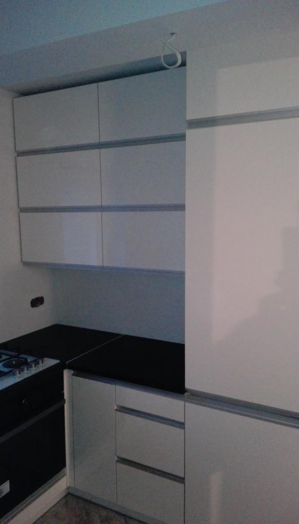 Białe, błyszczące szafki kuchenne z czarnym blatem, widoczna płyta grzewcza, minimalistyczny design, kabel zwisający z sufitu.