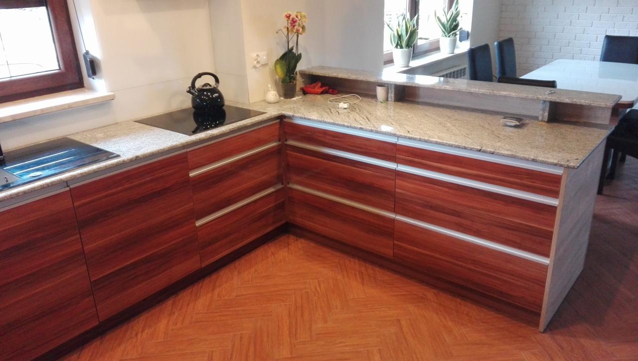 Kuchenna zabudowa z drewnopodobnymi frontami i blatem z jasnego granitu, widoczny czarny czajnik na płycie indukcyjnej, parkiet w jodełkę na podłodze.