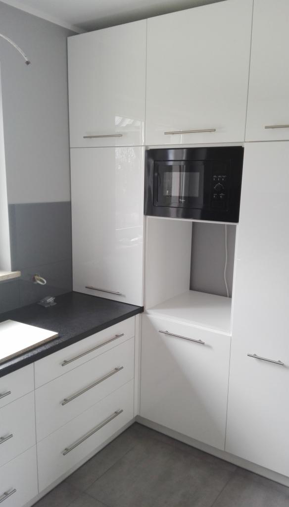 Biała, lakierowana zabudowa kuchenna z czarnym blatem i wbudowaną mikrofalówką, widok na narożnik szafek.