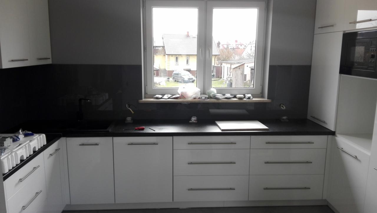 Białe szafki kuchenne z czarnym blatem, widoczne akcesoria montażowe na parapecie i narzędzia na blacie, okno z widokiem na zabudowę jednorodzinną.