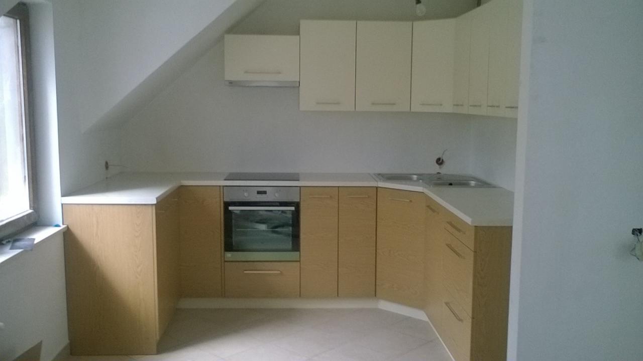 Jasna kuchnia w skosach, z drewnopodobnymi frontami szafek dolnych i białymi górnymi, biały blat, okno po lewej stronie, płytki podłogowe w neutralnym odcieniu.