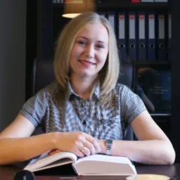 Uśmiechnięta blondynka w biurze, siedząca za biurkiem z otwartą książką, na palcu złoty pierścionek, za nią regał z segregatorami.