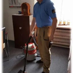 Pracownik w niebieskiej koszulce z logiem, odkurza dywan w biurze odkurzaczem przemysłowym, widoczne biurko z radiem i kalendarzem na ścianie.