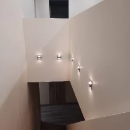 Wnętrze klatki schodowej z oświetleniem ściennym LED, widok z góry na schody i drzwi na podeście, minimalistyczny design.