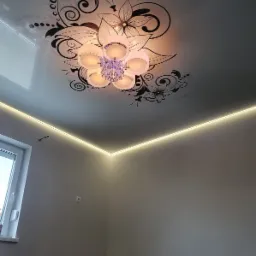 Biały sufit napinany z czarnym motywem kwiatowym i ozdobnym żyrandolem, oświetlony ledowym paskiem wzdłuż krawędzi ściany.