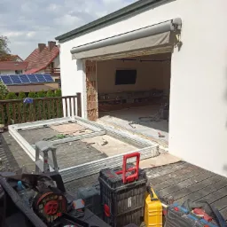 Demontaż starego okna na tarasie z drewnianą podłogą, widoczne narzędzia i skrzynki narzędziowe, w tle domy z panelami słonecznymi.