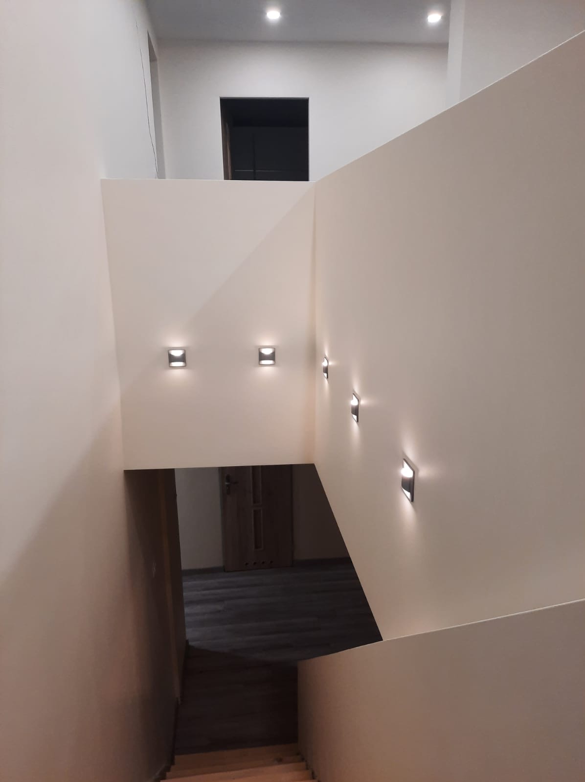 Wnętrze klatki schodowej z oświetleniem ściennym LED, widok z góry na schody i drzwi na podeście, minimalistyczny design.