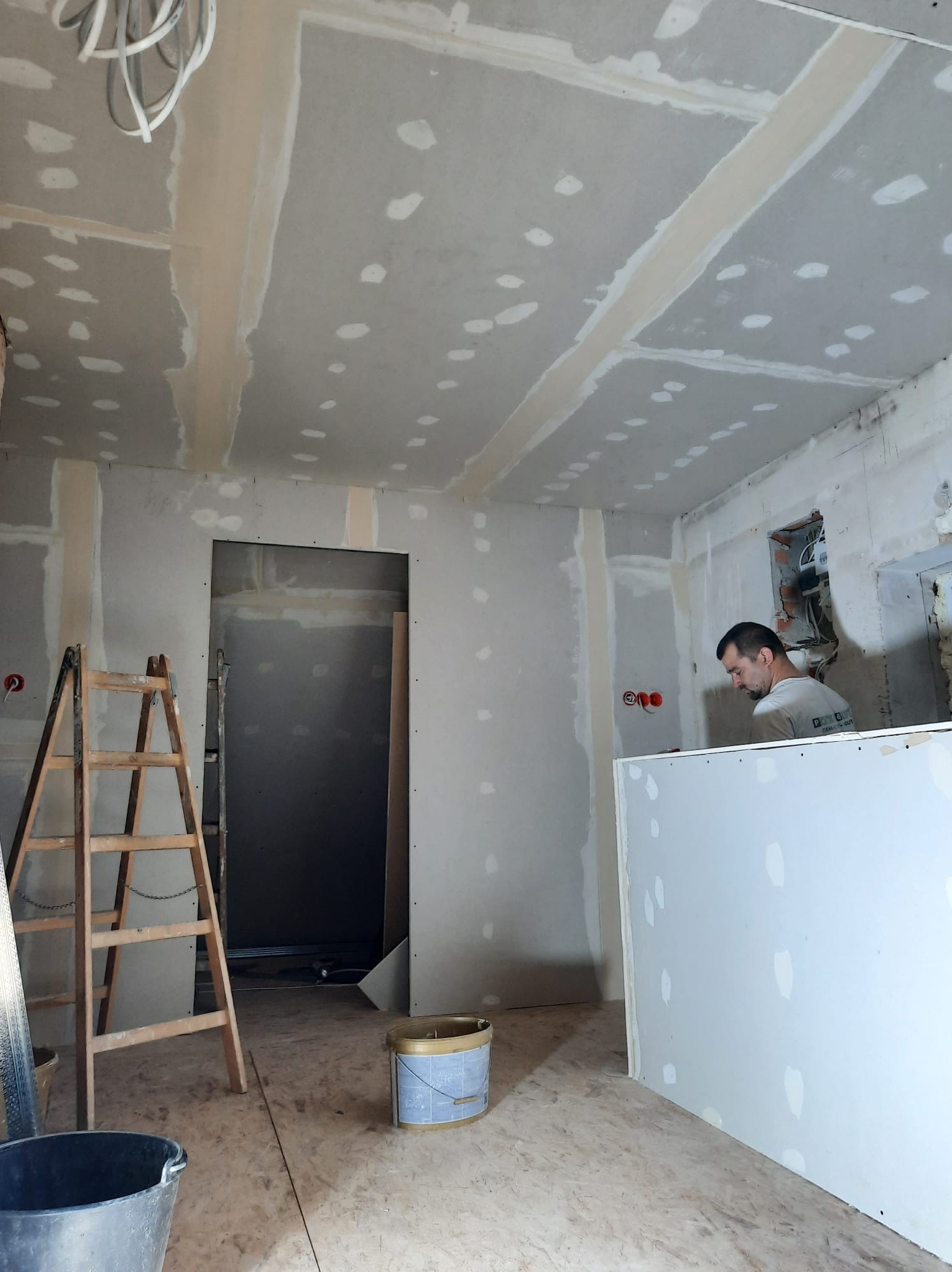 Prace wykończeniowe wnętrz: montaż płyt karton gips na ścianach i suficie, widoczna drabina, wiadro i pracownik w trakcie szpachlowania łączeń.