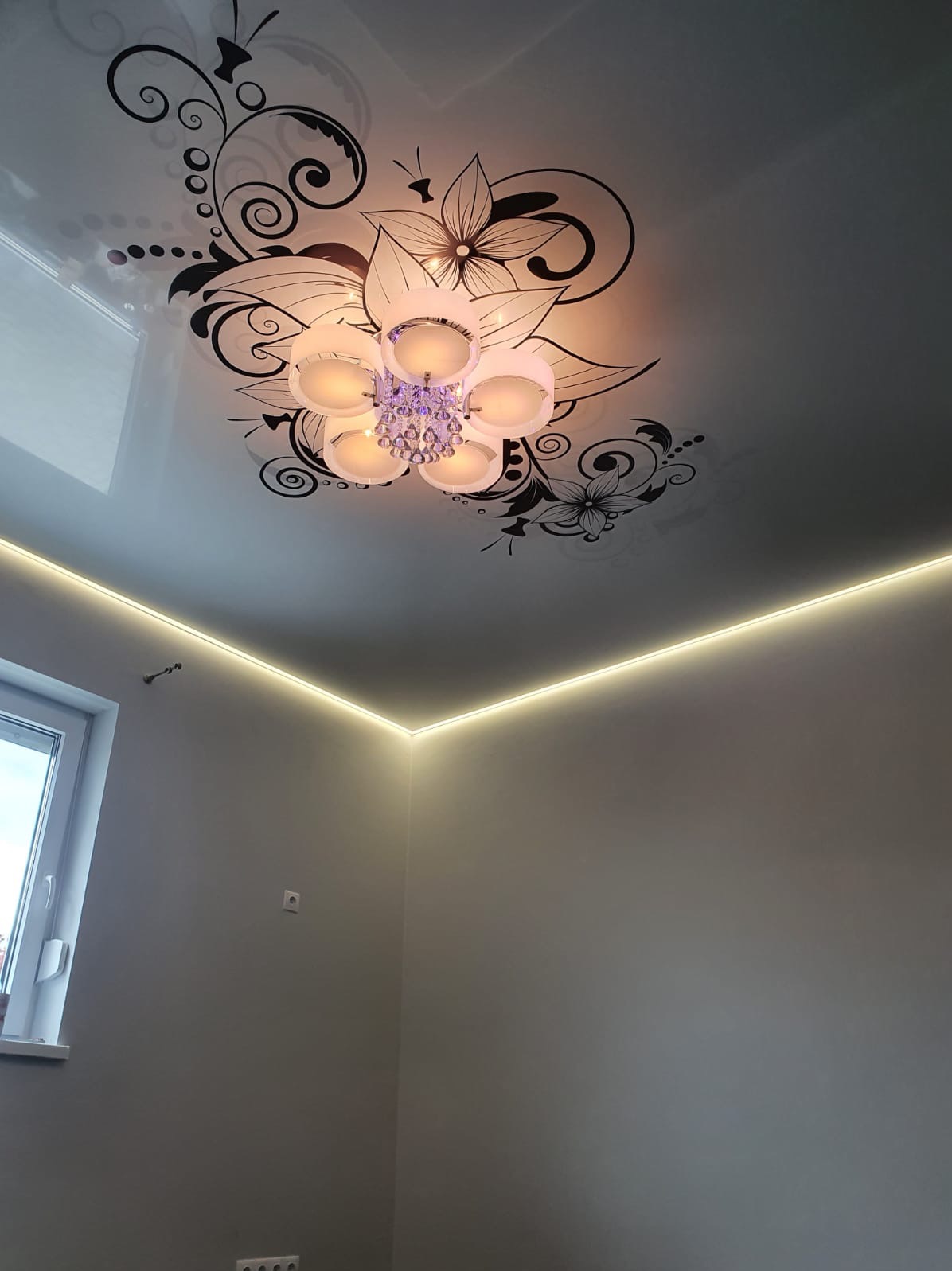 Biały sufit napinany z czarnym motywem kwiatowym i ozdobnym żyrandolem, oświetlony ledowym paskiem wzdłuż krawędzi ściany.