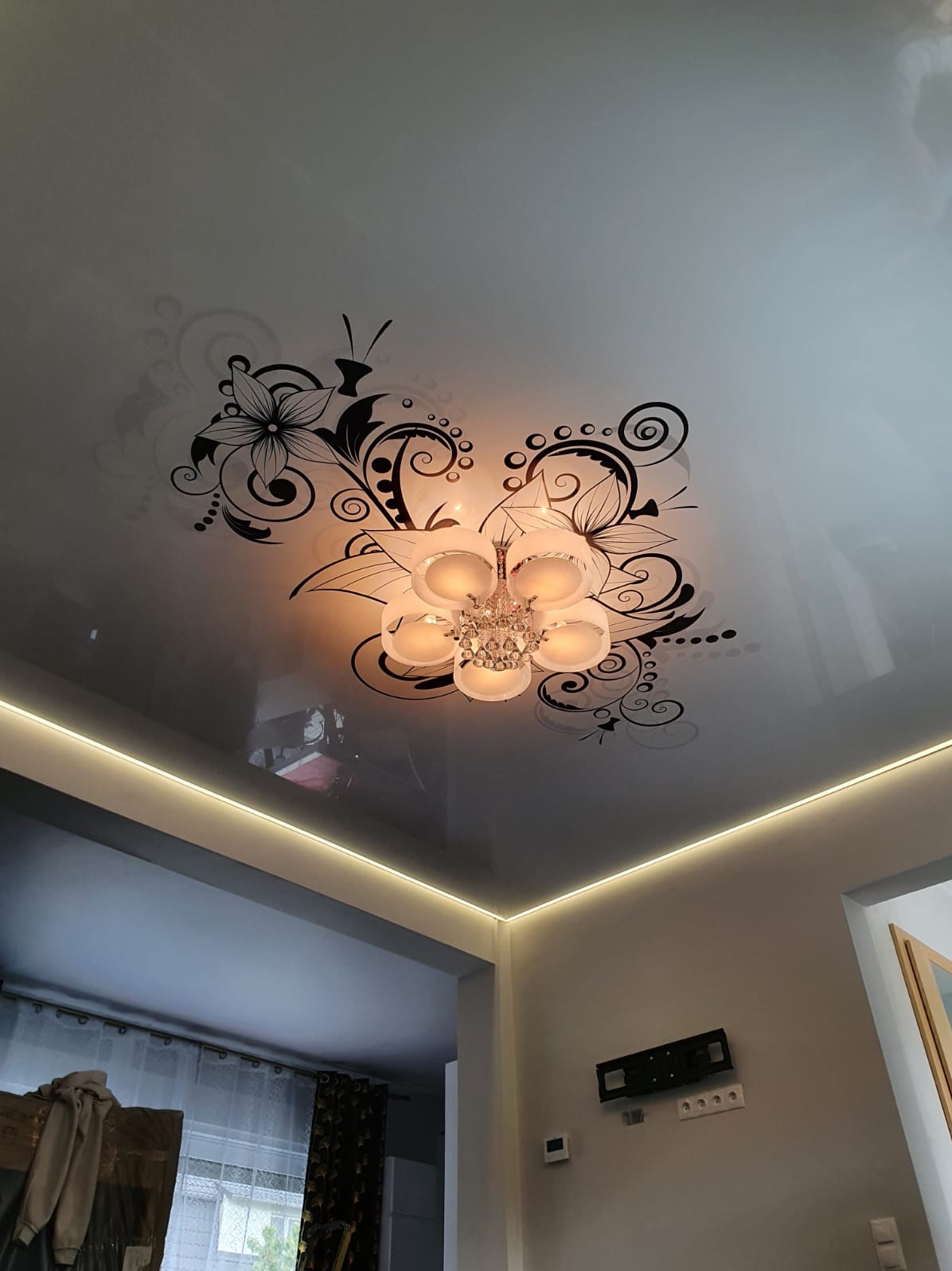 Biały, błyszczący sufit z czarnymi, kwiatowymi ornamentami i kryształowym żyrandolem w centrum, oświetlenie LED wzdłuż krawędzi sufitu.