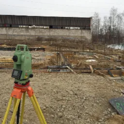 Zielony teodolit Leica na pomarańczowych nóżkach, ustawiony na placu budowy z widocznymi fundamentami i zbrojeniami, w tle budynek przemysłowy i drzewa bez liści.