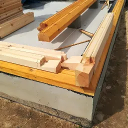 Fragment fundamentu z częściowo wzniesioną konstrukcją z drewnianych belek z wycięciami na łączeniach, przygotowane elementy konstrukcyjne ułożone obok.