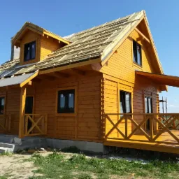 Żółty dom z drewnianych bali w trakcie budowy, z widocznym częściowo ułożonym dachem i drewnianą werandą z balustradą w kształcie litery X.
