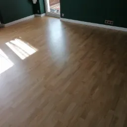 Jasna, drewniana podłoga ułożona w jodełkę, z widocznym fragmentem ściany w kolorze butelkowej zieleni i promieniami słonecznymi wpadającymi przez okno.