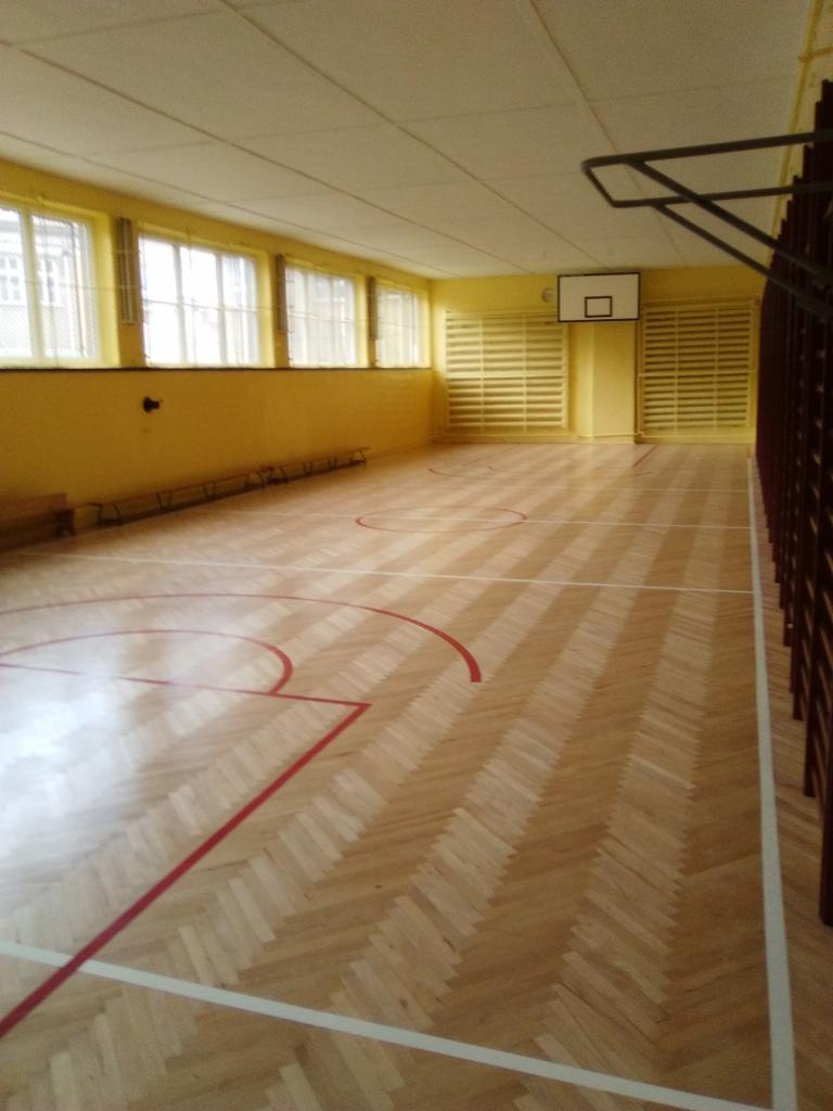 Sala gimnastyczna z parkietem ułożonym w jodełkę, widoczne linie boiska, kosz do koszykówki i okna z kratami.