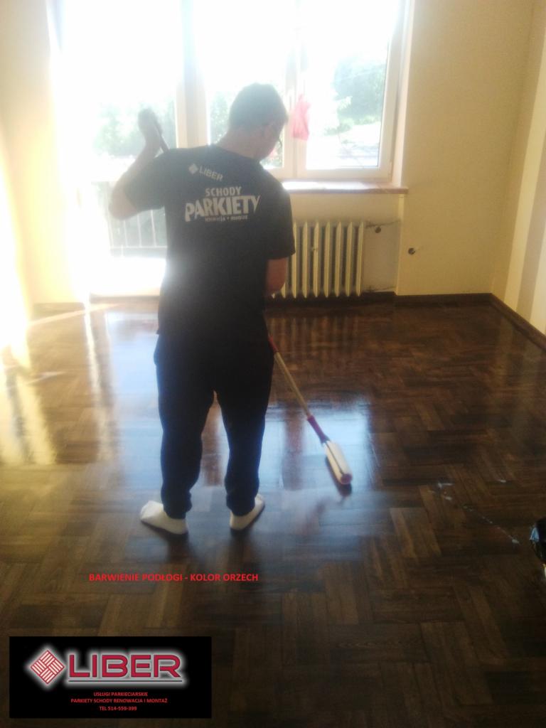 Parkiet w jodełkę po barwieniu na kolor orzechowy, pracownik w firmowej koszulce rozprowadza preparat, widoczne odbicie światła na świeżo pokrytej powierzchni.