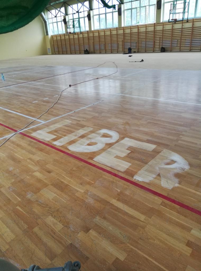 Sala gimnastyczna z drewnianą podłogą w trakcie renowacji, widoczne częściowo odnowione logo 'LIBER' oraz przewód od maszyny do obróbki podłóg.