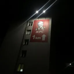 Duży, podświetlony nocą baner reklamowy KFC umieszczony na szczycie budynku mieszkalnego z oknami, wskazujący odległość 1 km w górę.